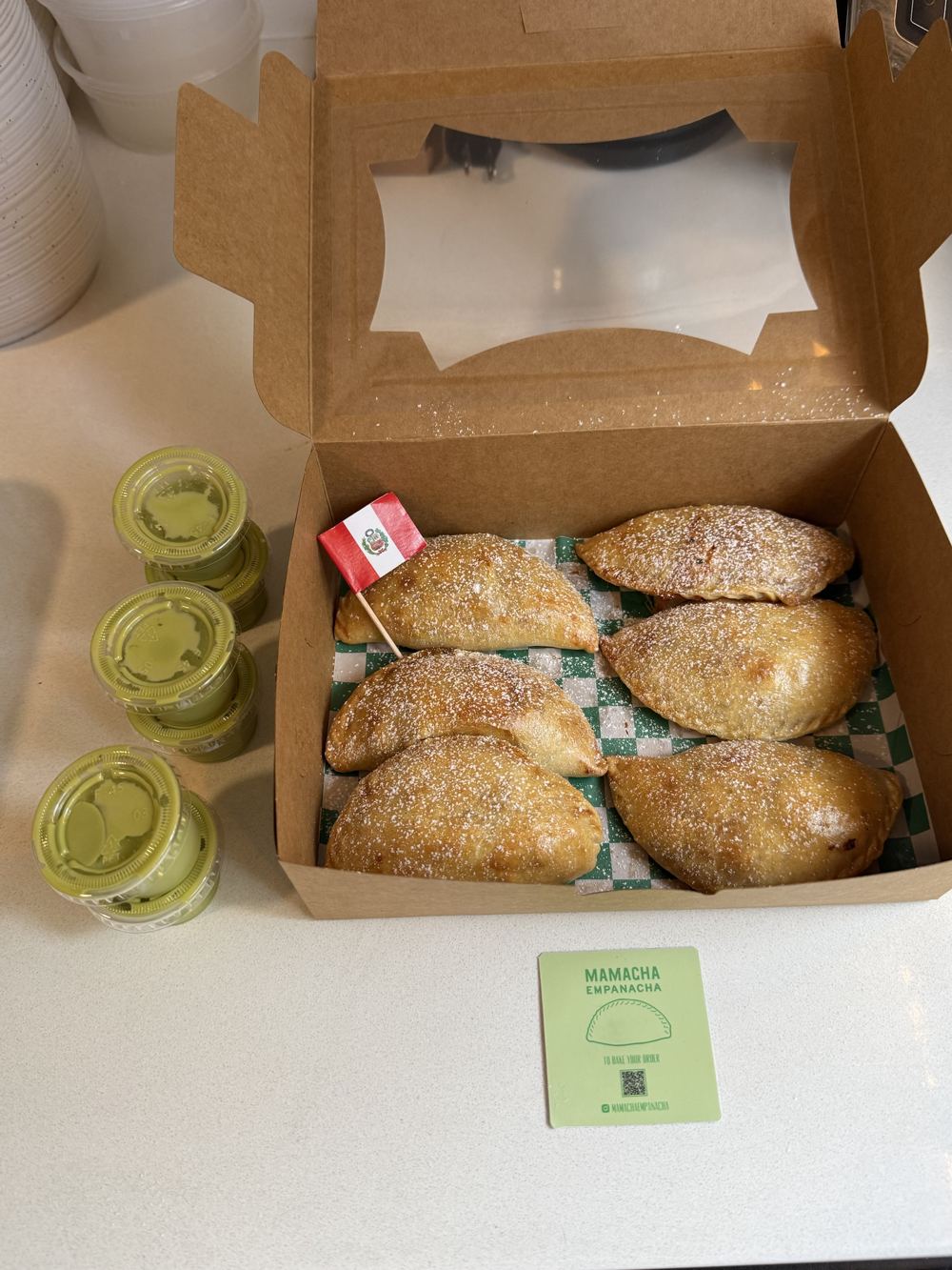 An open box of six empanaditas with a Peruvian flag pick, next to cups of crema
